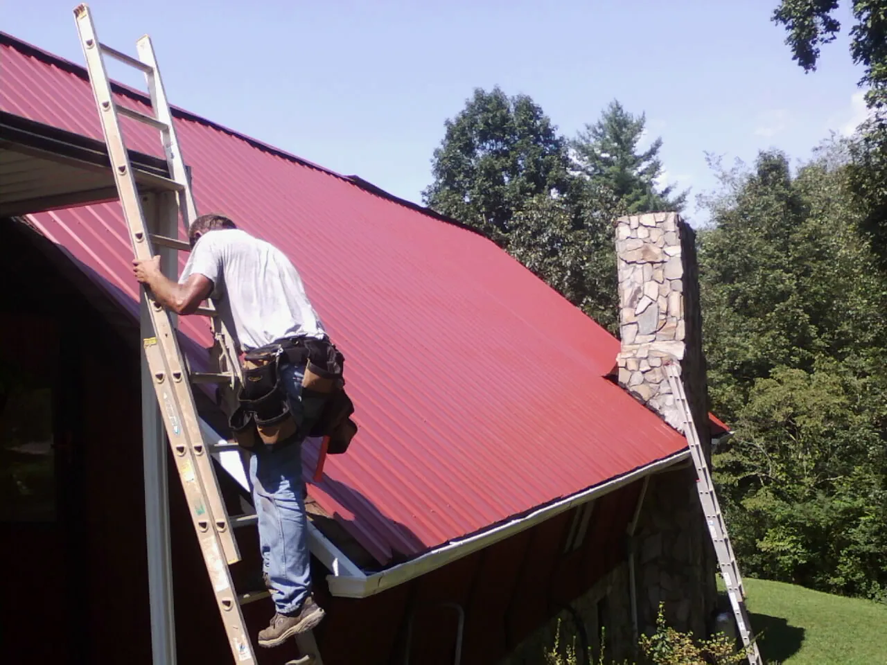 Roofer on a ladder beside a red metal roof and stone chimney — Soffit & Fascia Repair by Roofing Brothers in Mcclusky